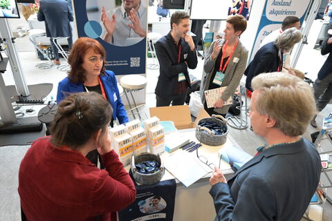 Aufnahme einer Messesituation von oben. Zwei Frauen stehen an einem mit Material befüllten Tisch und interagieren mit einem Messebesucher. Im Hintergrund unterhalten sich mehrere Personengruppen