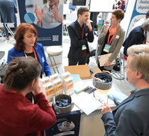 Aufnahme einer Messesituation von oben. Zwei Frauen stehen an einem mit Material befüllten Tisch und interagieren mit einem Messebesucher. Im Hintergrund unterhalten sich mehrere Personengruppen