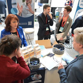 Aufnahme einer Messesituation von oben. Zwei Frauen stehen an einem mit Material befüllten Tisch und interagieren mit einem Messebesucher. Im Hintergrund unterhalten sich mehrere Personengruppen