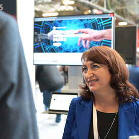 Eine vhs-Vertreterin mittleren Alters in royalblauem Sakko steht vor einem Bildschirm und unterhält sich mit einem Messegast