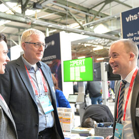 Zwei vhs-Vertreter und eine vhs-Vertreterin unterhalten sich angeregt. Im Hintergrund sieht man ein vhs-Banner und einen grün leuchtenden Bildschirm mit XPert-Logo