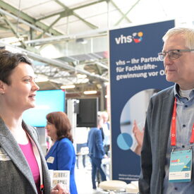 Eine vhs-Vertreterin mittleren Alters und ein älterer vhs-Vertreter im Gespräch vor dem vhs-Messebanner