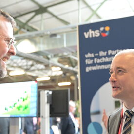 Zwei vhs-Vertreter mittleren Alters im Gespräch vor dem vhs-Messebanner