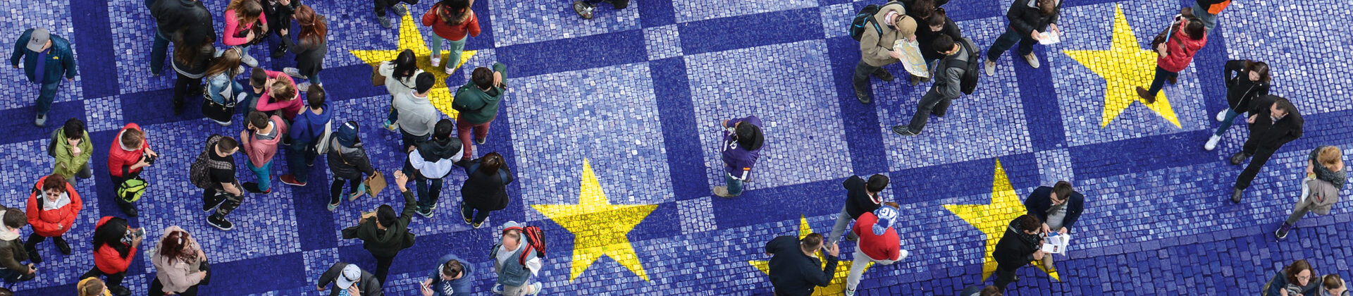 Vogelperspektive auf einen öffentlichen Platz, über den ganz viele Menschen laufen. Der Platz ist im Blau der europäischen Union eingefärbt und darauf sind gelbe Sterne zu sehen.
