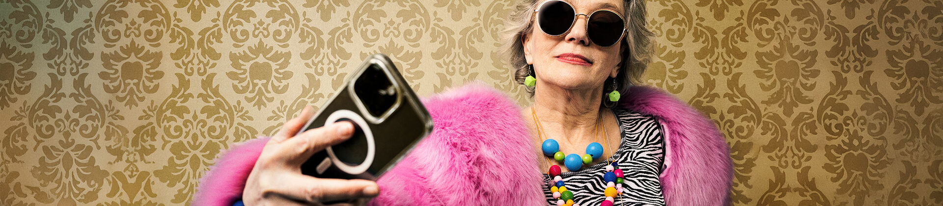 Eine ältere Damen in einem Rosa Mantel und mit großem bunten Schmuck macht ein Selfie.