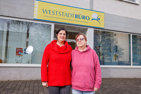 Eine Frau mittleren Alters und eine ältere Frau stehen zusammen vor einem Gebäude mit der Aufschrift "Hanau Weststadtbüro" und lächeln in die Kamera