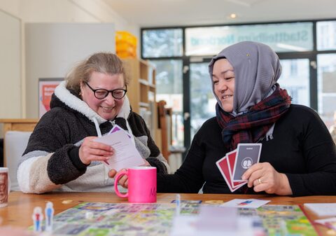 Zwei ältere Frauen sitzen lächelnd an einem großen Tisch und arbeiten gemeinsam mit Lernkarten. Auf dem Tisch liegt ein Brettspiel, davor steht ein Kaffeebecher