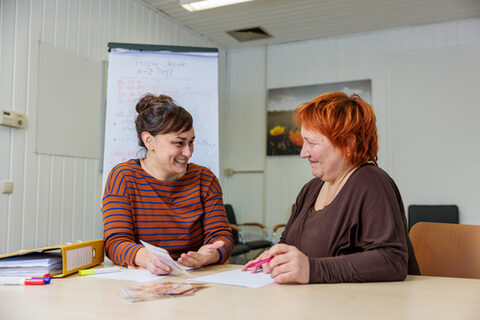 Zwei Frauen in mittlerem Alter sitzen mit Papier und Stiften nebeneinander an einem Schreibtisch und bearbeiten zusammen Lernkarten.