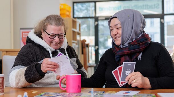 Zwei Frauen mittleren Alters, von denen eine ein Kopftuch trägt, sitzen lächelnd an einem großen Schreibtisch und arbeiten mit Lernkarten