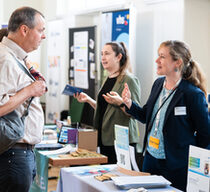 Zwei jüngere Frauen in Businessoutfits stehen hinter Tischen mit Informationsmaterial und sprechen mit einem Mann und einer Frau, im Hintergrund sieht man eine Europaflagge.