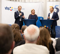 Drei Personen stehen an Stehstischen und sprechen zu einem Publikum. Zwei von ihnen halten ein Mikrofon in der Hand.