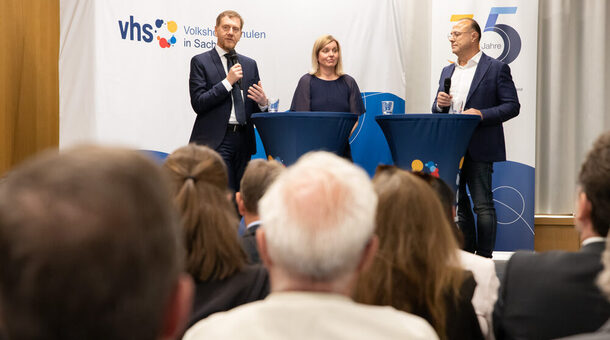 Drei Personen stehen an Stehstischen und sprechen zu einem Publikum. Zwei von ihnen halten ein Mikrofon in der Hand.