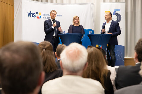 Drei Personen stehen einem Publikum zugewandt. Einer von ihnen spricht in ein Mikrofon.