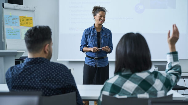 Eine junge Fraun in einem Jeanshemd steht vor einer Klasse und unterichtet