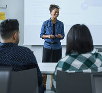 Eine junge Fraun in einem Jeanshemd steht vor einer Klasse und unterichtet