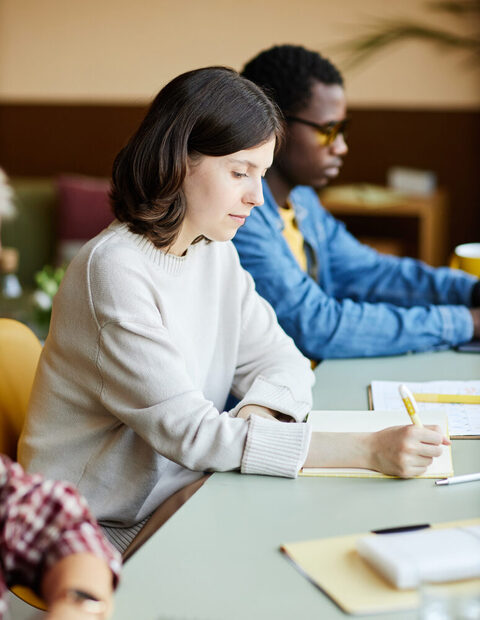 Drei Menschen sitzen an einem langen Tisch. In der Mitte eine junge Frau, die mit ernstem Blick auf ein Notizbuch schaut und etwas hineinschreibt.