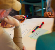 Vier Personen sitzen an einem runden Tisch und beschriften bunte Papierkarten. Auf einer Karte steht: "Was ist für dich Freiheit?"