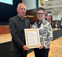 Ein älterer Mann im schwarzen Anzug und eine ältere Frau mit blaugemusterter Jacke halten lächelnd eine gerahmte Urkunde in die Kamera.