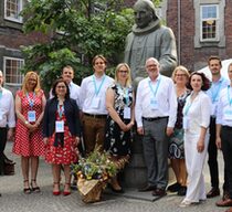 Fünf Frauen und sechs Männer in förmlicher Kleidung stehen als Gruppe auf einem Platz an einem Denkmal zusammen und lächeln in die Kamera.