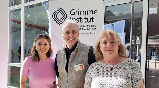 Zwei sommerlich gekleidete Frauen und ein Mann stehen zusammen vor einem Gebäude und lächeln in die Kamera.