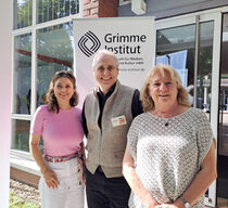 Zwei sommerlich gekleidete Frauen und ein Mann stehen zusammen vor einem Gebäude und lächeln in die Kamera.