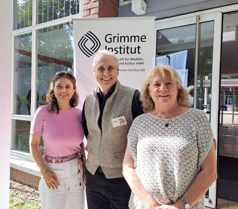 Zwei sommerlich gekleidete Frauen und ein Mann stehen zusammen vor einem Gebäude und lächeln in die Kamera.