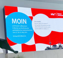 Screen mit Willkommensmotiv: rot-weiße Bremer Flagge im Hintergrund, ein großer hellblauer Kreis mit der Aufschrift: "Moin!" und einer kleinen Skyline-Grafik.
