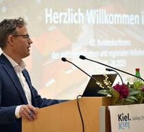 Ein Mann im Anzug steht an einem Rednerpult, im Hintergrund ein Wilkommensbildschirm.