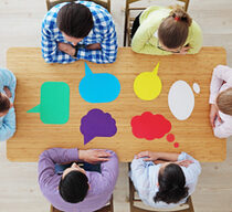 Blick aus der Vogelperspektive: Sechs Personen sitzen an einem Holztisch. Vor ihnen liegen jeweils Papiersprechblasen in unterschiedlichen Formen und Farben.