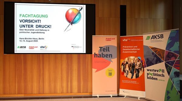 Ein Blick einer Leinwand auf die eine Folie mit dem Titel der Fachtagung "Vorsicht unter Druck!" projiziert ist. Daneben stehen drei Roll-Ups der Veranstalter Arbeit und Leben, Deutscher Volkshochschul-VErband und AKSB