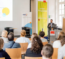 Eine Gruppe Personen sitzt auf Stühlen mit dem Rücken zur Kamera. Vorne am Rednerpult steht ein Mann und spricht zu der Gruppe.