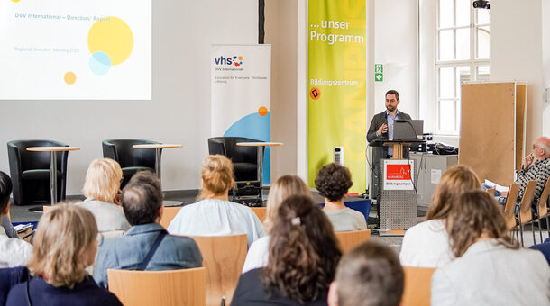 Eine Gruppe Personen sitzt auf Stühlen mit dem Rücken zur Kamera. Vorne am Rednerpult steht ein Mann und spricht zu der Gruppe.
