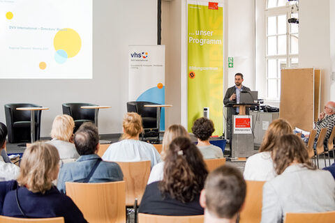 Eine Gruppe Personen sitzt auf Stühlen mit dem Rücken zur Kamera. Vorne am Rednerpult steht ein Mann und spricht zu der Gruppe.