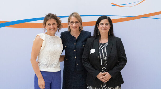 Drei festlich gekleidete Frauen stehen vor einer blau-orangenen Logowand und lächeln in die Kamera.