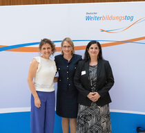 Drei festlich gekleidete Frauen stehen vor einer blau-orangenen Logowand und lächeln in die Kamera.