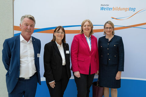 Drei festlich gekleidete Frauen und ein Mann stehen vor einer blau-orangenen Logowand und lächeln in die Kamera.