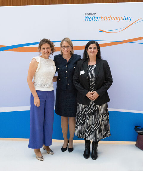 Drei festlich gekleidete Frauen stehen vor einer blau-orangenen Logowand und lächeln in die Kamera.