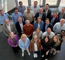 Eine Gruppe von Männern und Frauen steht zusammen in einem Flur und schaut lächelnd nach oben in die Kamera.