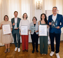 Vier Frauen und zwei Männer in festlichen Outfits stehen in einem Saal und halten Preise und Urkunden hoch.