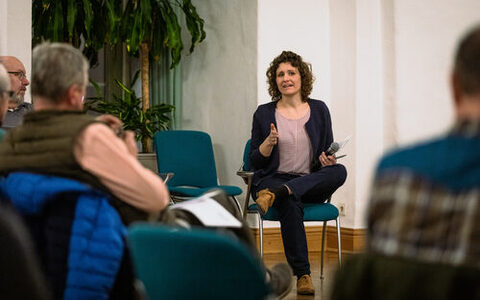 Eine Frau sitzt auf einem Stuhl einer Gruppe Personen zugewandt und spricht zu ihnen.