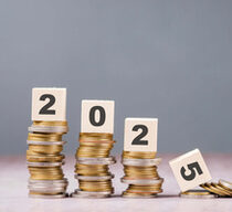 Das Bild zeigt vier Stapel Münzen, die von links nach rechts an Höhe abnehmen. Auf jedem Stapel liegt ein Holzklotz mit einer aufgedruckten Zahl. Von links nach rechts stehen die Zahlen 2, 0, 2 und 5, was zusammen die Jahreszahl 2025 ergibt. Der Stapel mit der Zahl 5 ist deutlich kleiner und der Holzklotz liegt leicht schräg auf den Münzen, als ob er gerade heruntergefallen wäre. Der Hintergrund ist grau und neutral gehalten.