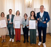 Vier Frauen und zwei Männer in festlichen Outfits stehen in einem Saal und halten Preise und Urkunden hoch.