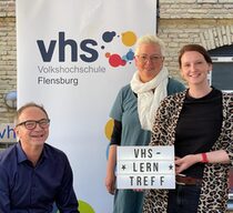 Drei Menschen stehen vor Schild der Volkshochschule Flensburg.