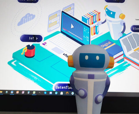 Eine Schaumstofffigur des Roboters aus der App "Stadt | Land | DatenFluss" ist vor einem Bildschirm mit dem Screenshot aus der App zu sehen